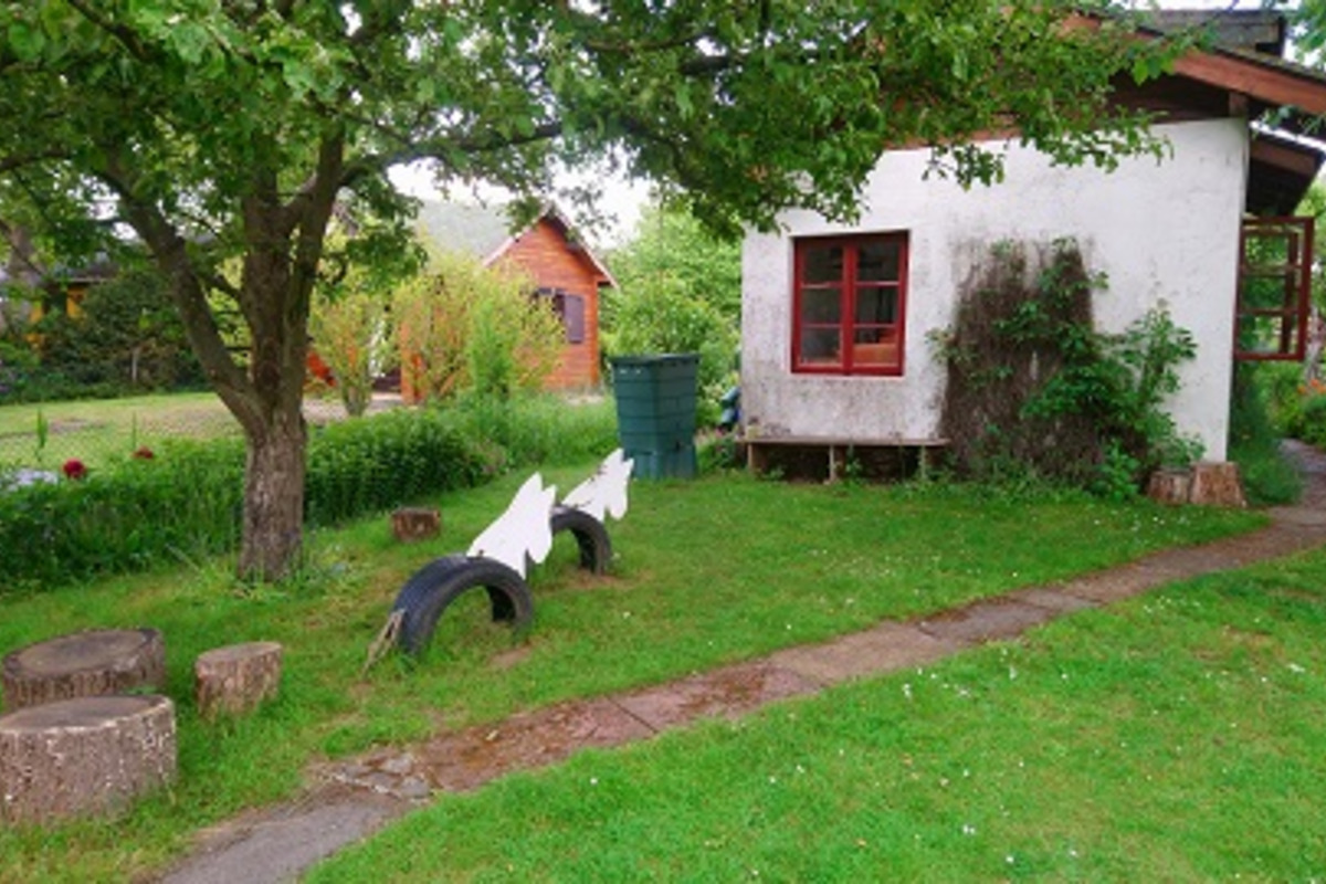 Ein kleines Wohnhaus mit roten Fensterrahmen steht in einem Garten. Vor dem Haus gibt es eine Rasenfläche mit Holzscheiten und zwei weißen, stilisierten Figuren. Im Hintergrund sind grüne Bäume und ein Mülltonne sichtbar. Der Weg zum Haus ist gepflastert.