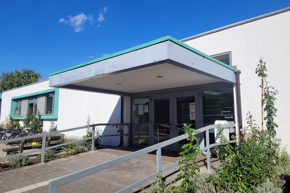 Eingang zu einem modernen, weißen Gebäude mit einem flachen Vordach und grünen Akzenten. Ein gepflasterter Weg führt zur Tür, flankiert von einigen Pflanzen. Auf der rechten Seite sind Fahrräder abgestellt. 