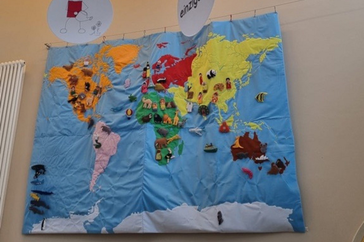 An einer Wand hängt eine farbenfrohe Weltkarte, die verschiedene Tiere und Symbole zeigt. Über der Karte sind mehrere runde Tafeln mit Sprüchen wie "Wir gestalten verlässliche Bindungen" und "Jeder ist einzigartig" angebracht, die eine Botschaft der Gleichwertigkeit und Gemeinschaft vermitteln.