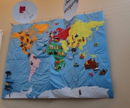 An einer Wand hängt eine farbenfrohe Weltkarte, die verschiedene Tiere und Symbole zeigt. Über der Karte sind mehrere runde Tafeln mit Sprüchen wie "Wir gestalten verlässliche Bindungen" und "Jeder ist einzigartig" angebracht, die eine Botschaft der Gleichwertigkeit und Gemeinschaft vermitteln.