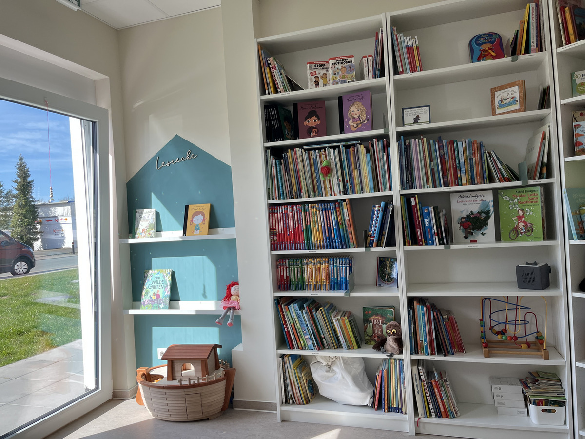 Ein helles Zimmer mit einer Bücherwand, die viele bunte Kinderbücher enthält. Links befindet sich ein Fenster mit Sicht auf eine grüne Wiese und ein Spielzeugboot auf dem Boden. An der Wand steht „lesecke“ und es gibt einige dekorative Elemente.