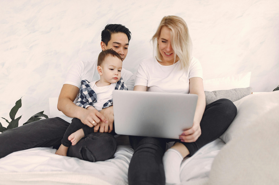 Eine Familie sitzt auf einem Bett und schaut gemeinsam auf einen Laptop. Ein Elternteil hält das Kleinkind auf dem Schoß, während der andere Elternteil den Laptop bedient. 