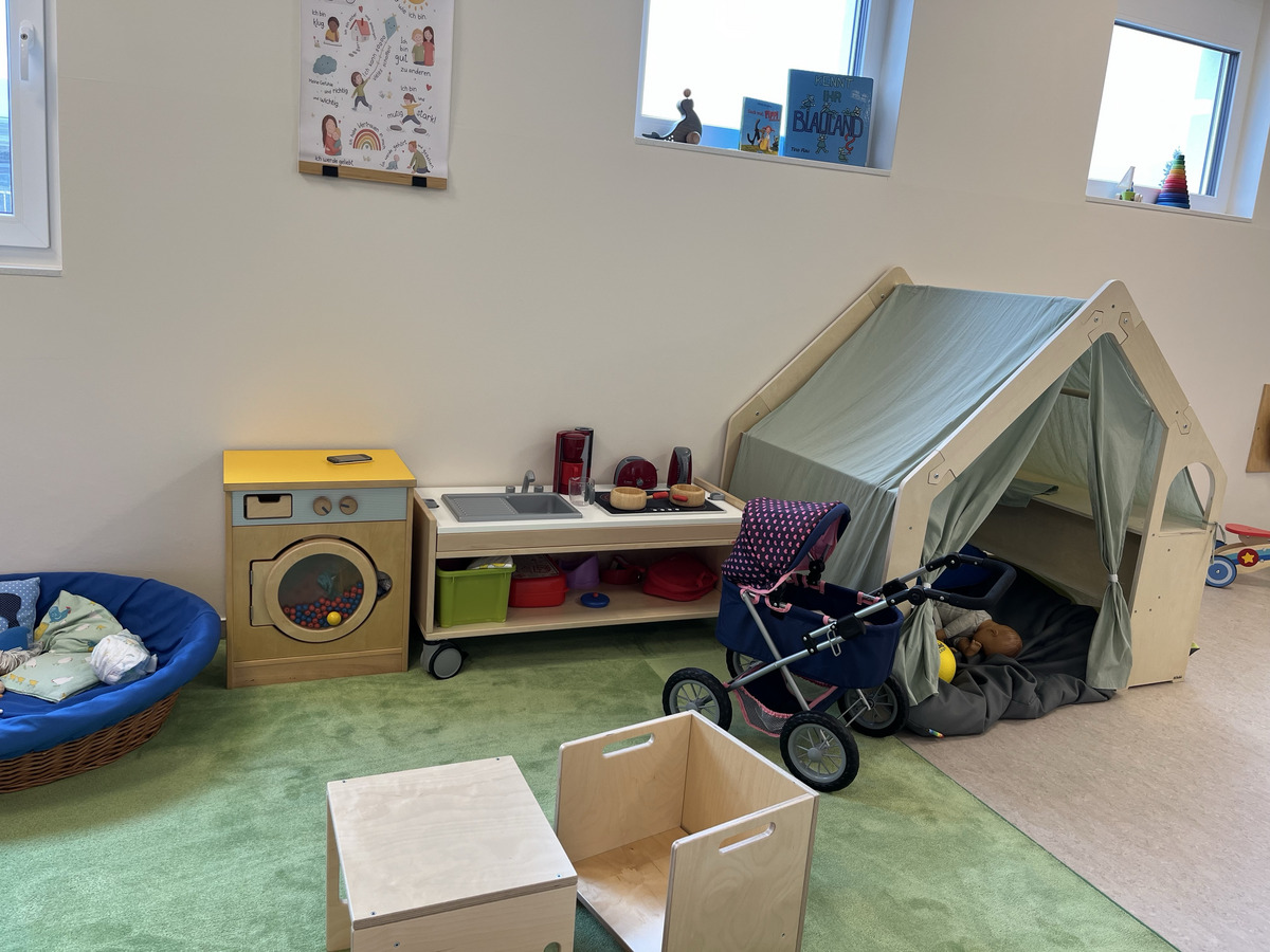 Ein Spielzimmer mit einem Spielzeugzelt in Form eines Hauses, einer Spielküche und einem Kinderwagen. Im Vordergrund befinden sich ein blauer Sessel und einige Holzspielzeuge auf grünem Teppich. Die Wände sind hell und kreativ dekoriert.