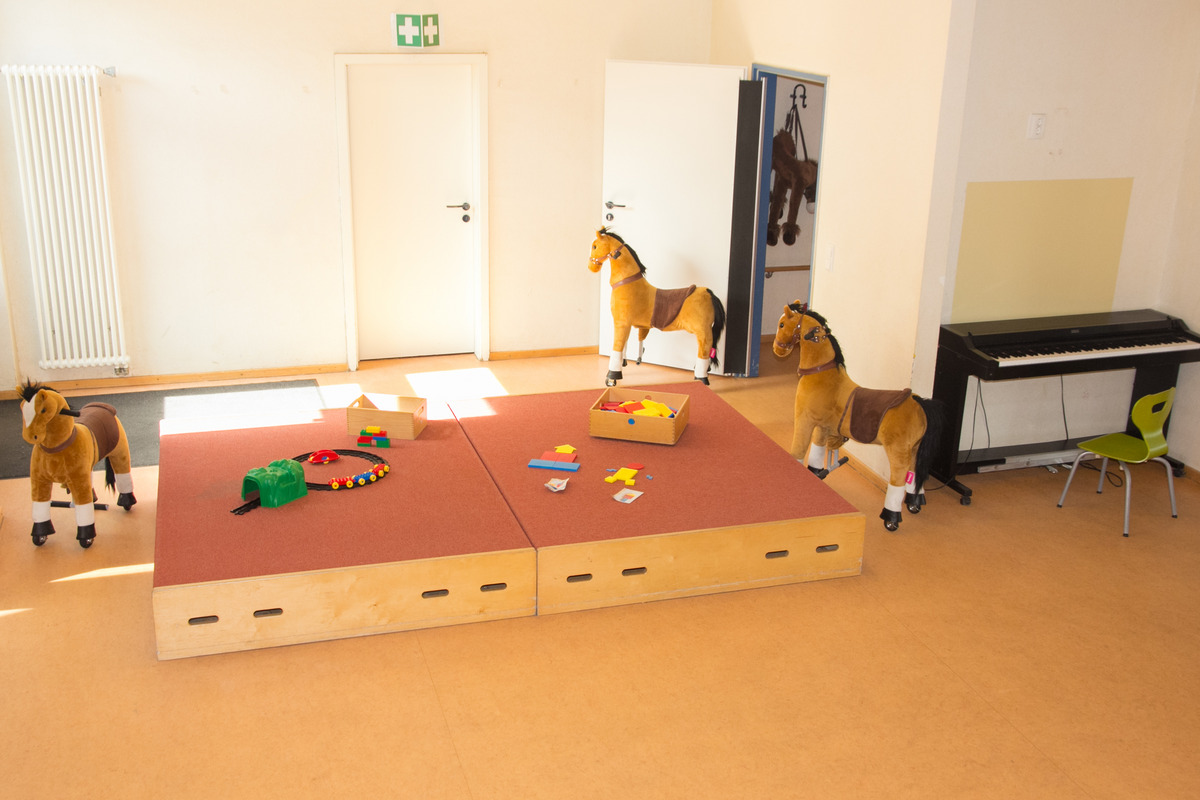 In einem hellen Raum stehen mehrere Plüschpferde auf einem roten Teppich. Auf einer erhöhten Holzunterlage liegen farbige Spielzeuge und Bauklötze. Im Hintergrund gibt es eine Wand mit einem Klavier und einer offenen Tür. Der Raum wirkt einladend und kindgerecht.