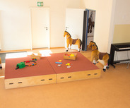 In einem hellen Raum stehen mehrere Plüschpferde auf einem roten Teppich. Auf einer erhöhten Holzunterlage liegen farbige Spielzeuge und Bauklötze. Im Hintergrund gibt es eine Wand mit einem Klavier und einer offenen Tür. Der Raum wirkt einladend und kindgerecht.