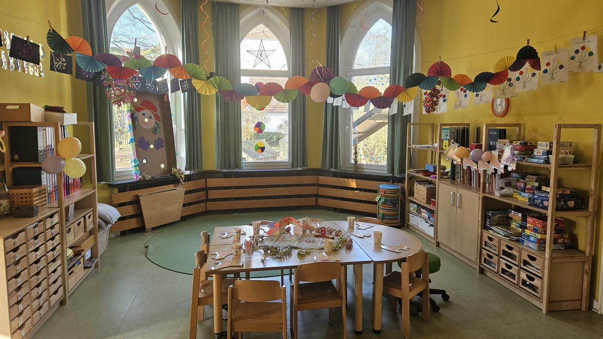 Im Bild ist ein bunter Gruppenraum zu sehen, der festlich dekoriert ist. Ein runder Tisch mit Stühlen steht in der Mitte, geschmückt mit Partyutensilien. An den Wänden hängen farbige Papiergirlanden und Kinderkunstwerke, während Fenster mit farbigen Dekorationen versehen sind.