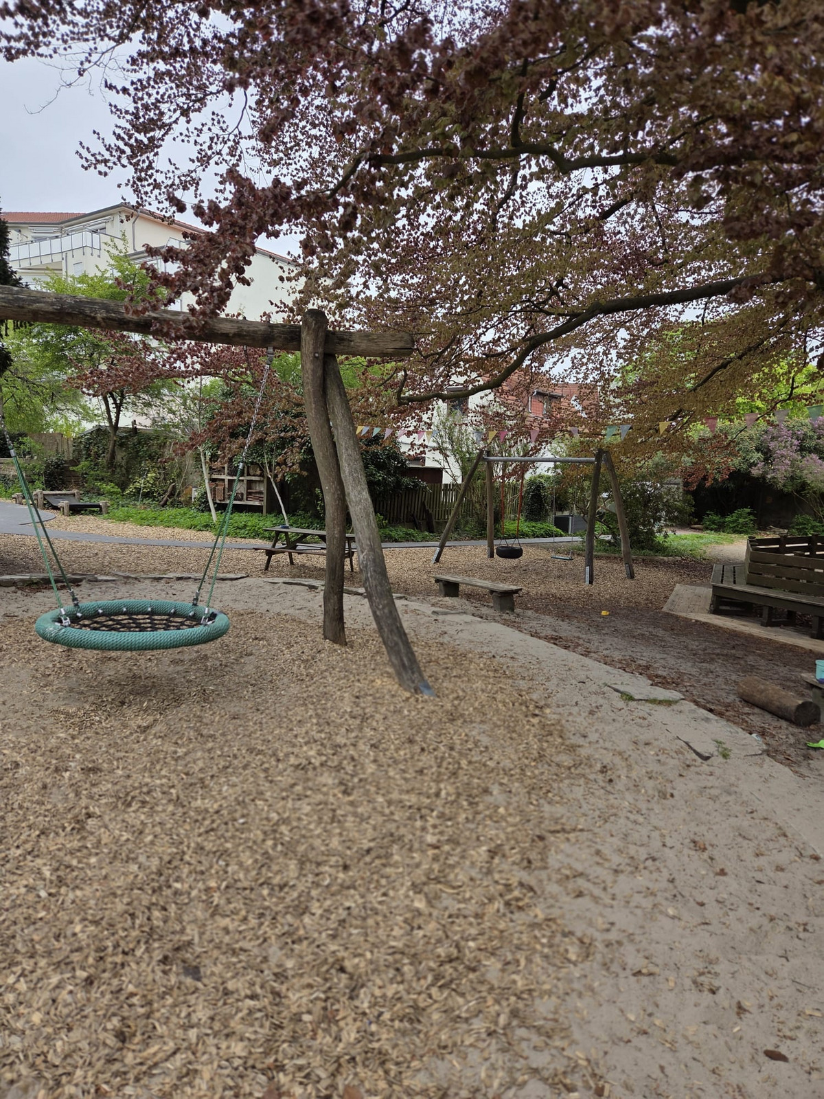 Ein Spielplatz mit einer großen, runden Schaukel und einer klassischen Schaukel aus Holz. Im Hintergrund sind Bänke und ein Spielbereich sichtbar. Über dem Spielplatz befinden sich Blütenzweige eines Baumes. Der Boden ist mit Holzspänen bedeckt.