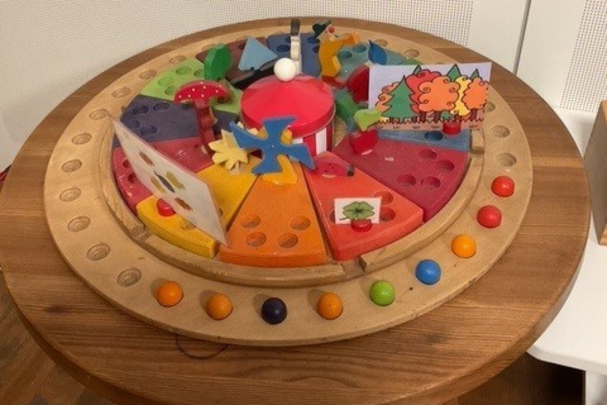 Ein runder Holztisch mit einem bunten, interaktiven Spielzeug. Das Spielzeug hat verschiedene farbige Segmente und enthält Figuren von Bäumen, einem Pilz und anderen Formen. 