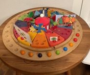 Ein runder Holztisch mit einem bunten, interaktiven Spielzeug. Das Spielzeug hat verschiedene farbige Segmente und enthält Figuren von Bäumen, einem Pilz und anderen Formen. 