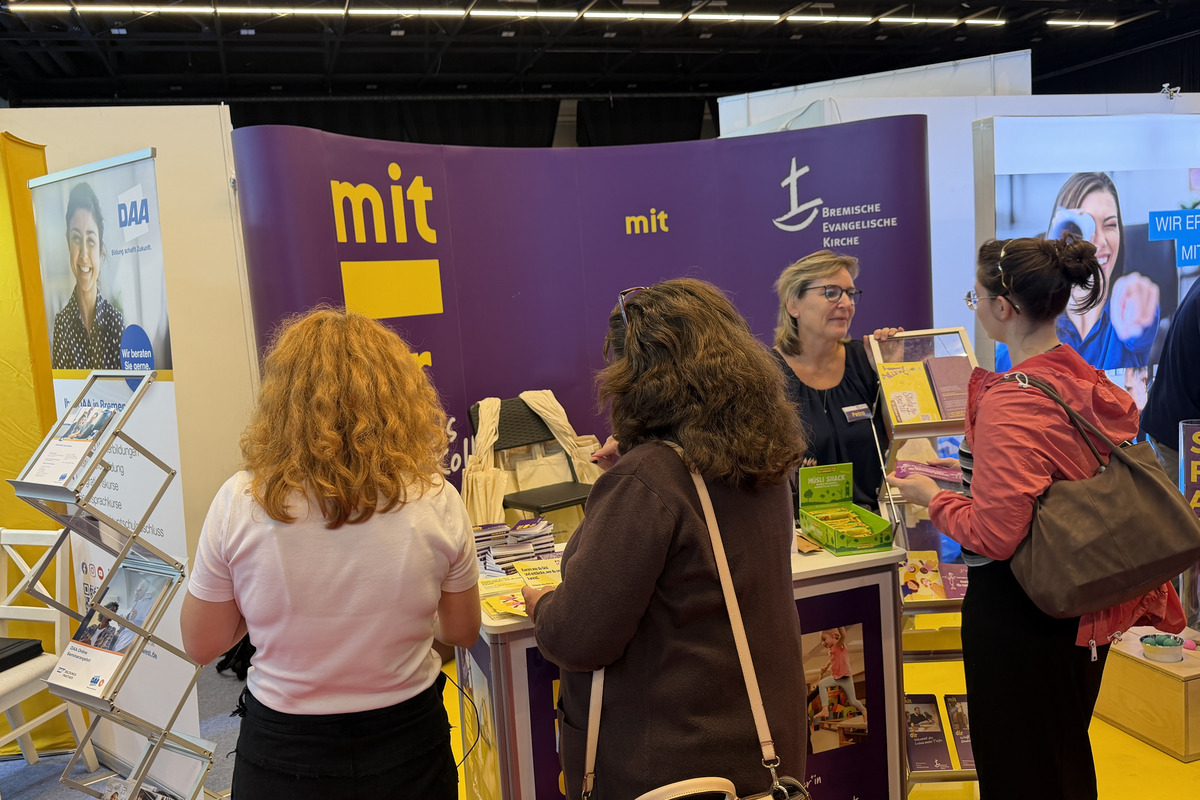 Mehrere Personen stehen an einem Informationsstand auf einer Messe. Der Stand präsentiert Informationsmaterialien, und eine Mitarbeiterin gibt Auskünfte. Im Hintergrund sind weitere Messestände zu sehen. Die Farben sind überwiegend lila und gelb.