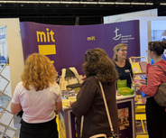Mehrere Personen stehen an einem Informationsstand auf einer Messe. Der Stand präsentiert Informationsmaterialien, und eine Mitarbeiterin gibt Auskünfte. Im Hintergrund sind weitere Messestände zu sehen. Die Farben sind überwiegend lila und gelb.