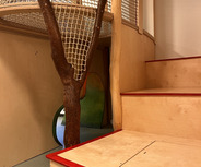 Ein Innenraum mit einem Spielbereich. Eine Holzkonstruktion mit Netz und zwei Baumstämmen dient als Kletterelement. Im Hintergrund sind farbige geschwungene Öffnungen sichtbar. Rechts führen Stufen hinauf zu einer Plattform.