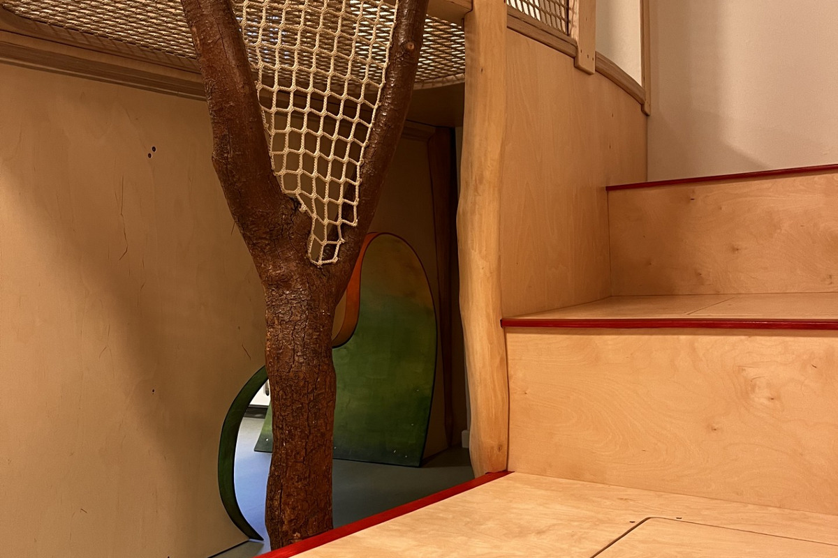Ein Innenraum mit einem Spielbereich. Eine Holzkonstruktion mit Netz und zwei Baumstämmen dient als Kletterelement. Im Hintergrund sind farbige geschwungene Öffnungen sichtbar. Rechts führen Stufen hinauf zu einer Plattform.