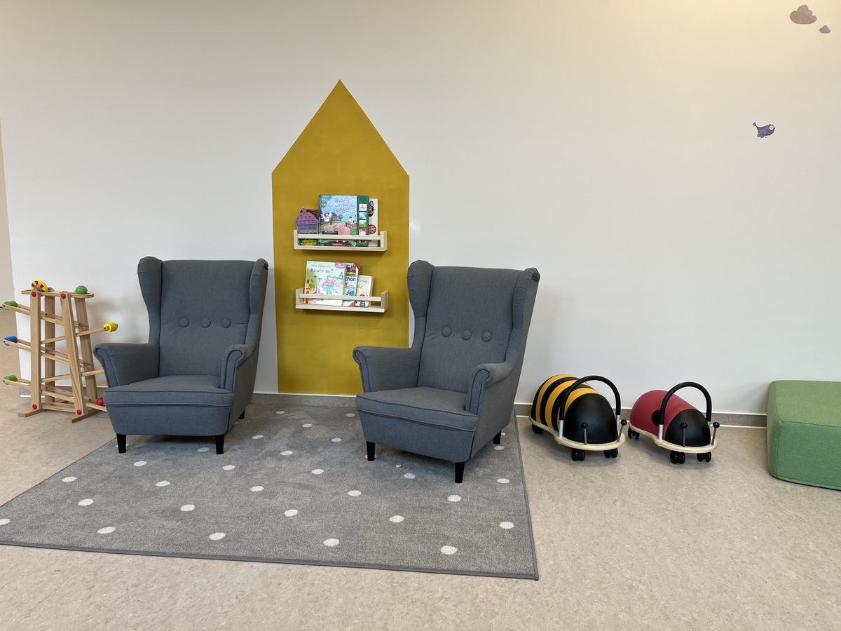 Der Raum zeigt zwei gemütliche graue Sessel auf einem Teppich mit weißen Punkten. An der Wand befindet sich ein Bücherregal mit bunten Kinderbüchern, flankiert von einer gelben Hausform. Auf dem Boden sind Spielzeuge in Form von Käfern.