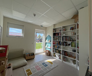Ein heller Raum mit einem Sofa, Spielzeugregalen und einer Bücherwand. Auf dem Boden liegt ein Teppich mit Zahlen. An der Wand sind Bilder und ein Regal mit Spielzeug. Durch ein Fenster ist eine grüne Wiese sichtbar.