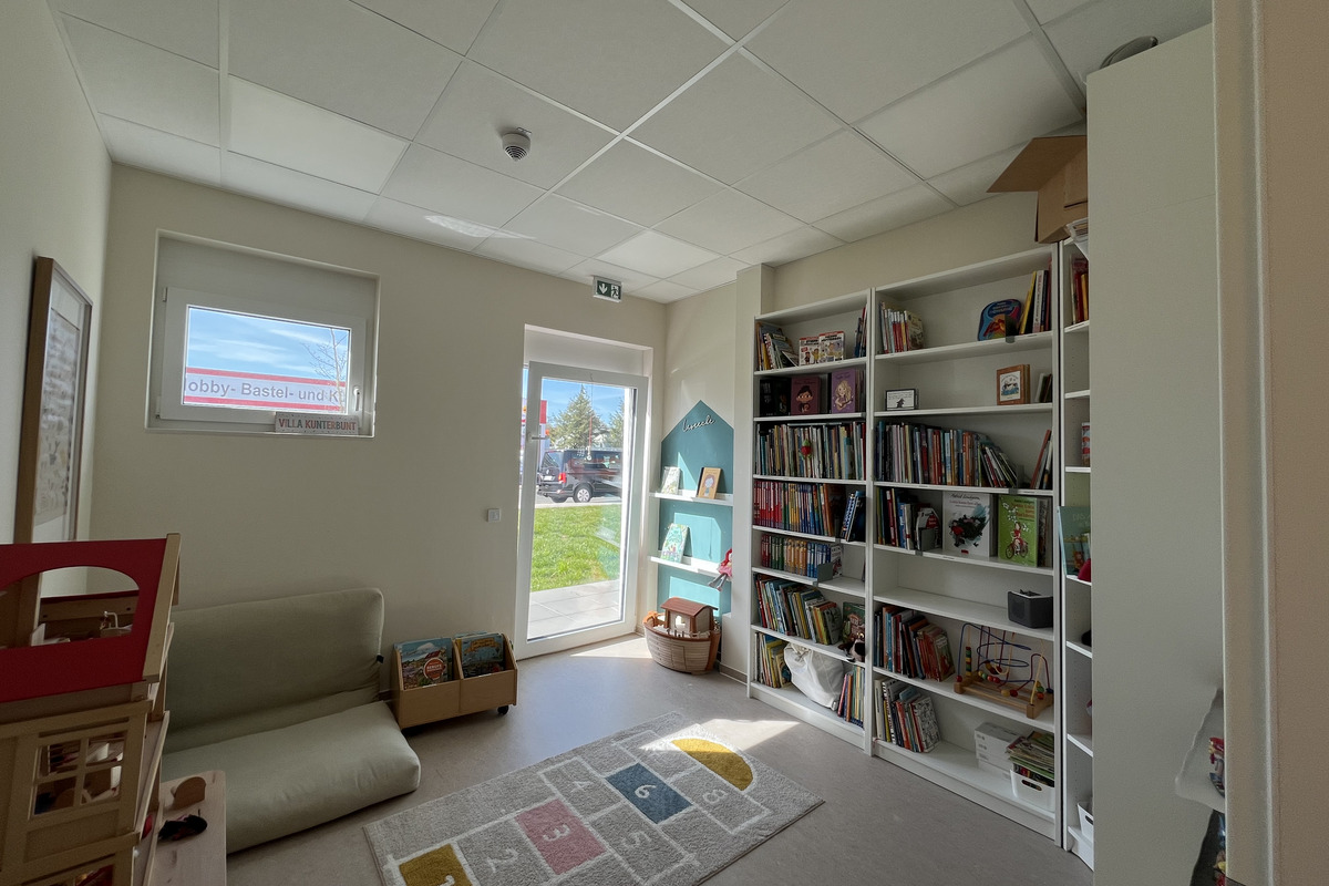 Ein heller Raum mit einem Sofa, Spielzeugregalen und einer Bücherwand. Auf dem Boden liegt ein Teppich mit Zahlen. An der Wand sind Bilder und ein Regal mit Spielzeug. Durch ein Fenster ist eine grüne Wiese sichtbar.