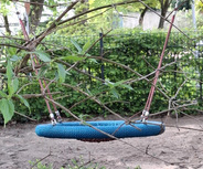 Eine blaue Nestschaukel hängt an einem Baum, umgeben von grünen Blättern und Ästen. Der Boden besteht aus Sand, und im Hintergrund sind Sträucher sichtbar. Die Szenerie vermittelt eine ruhige, natürliche Atmosphäre im Freien.