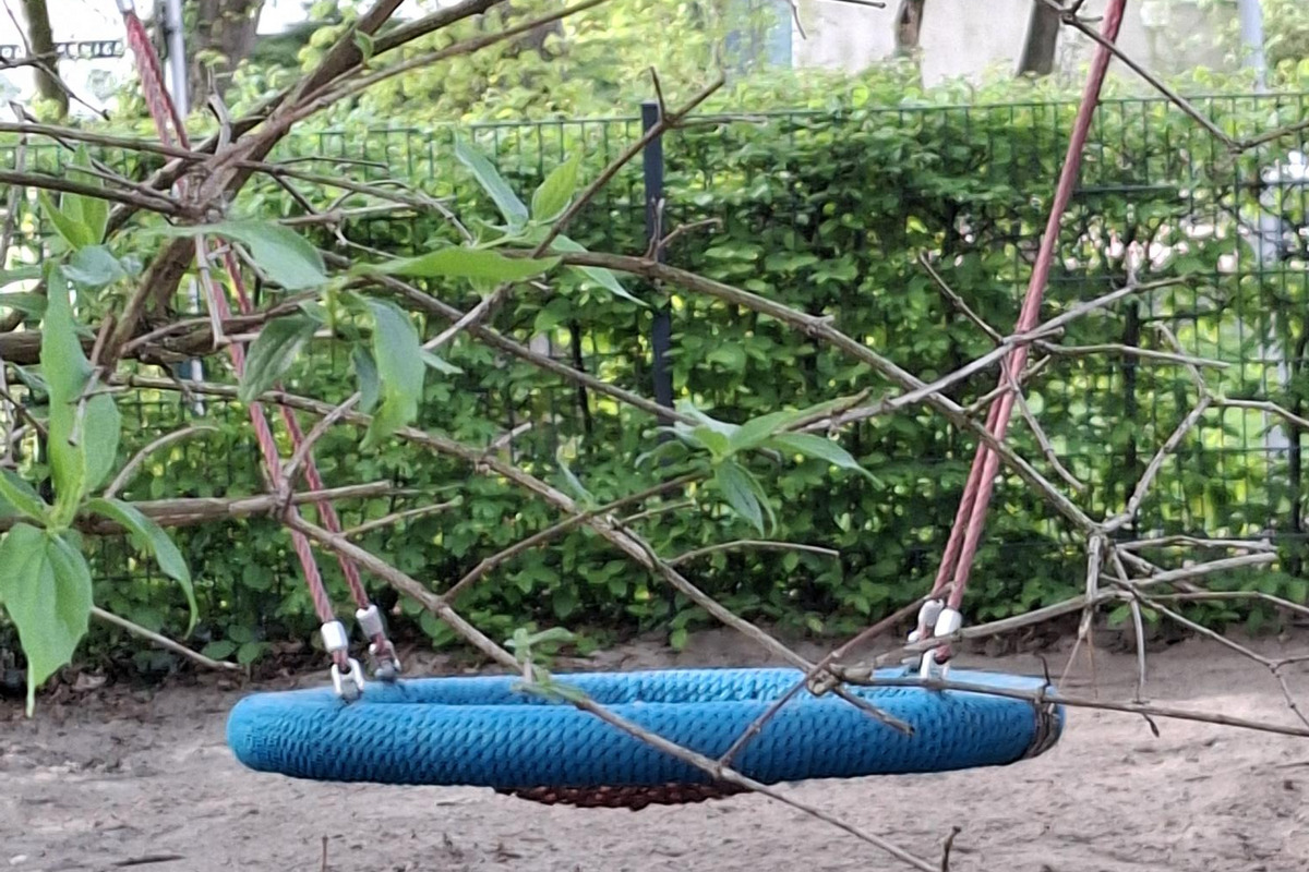 Eine blaue Nestschaukel hängt an einem Baum, umgeben von grünen Blättern und Ästen. Der Boden besteht aus Sand, und im Hintergrund sind Sträucher sichtbar. Die Szenerie vermittelt eine ruhige, natürliche Atmosphäre im Freien.
