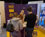 Ein Messestand mit einem großen Banner, auf dem "mit dir" steht. Menschen stehen zusammen, während sie Informationen austauschen. Im Hintergrund sind weitere Stände mit Werbematerialien zu sehen.