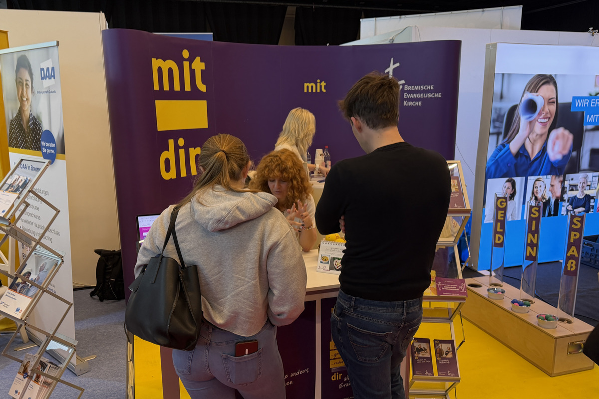 Ein Messestand mit einem großen Banner, auf dem "mit dir" steht. Menschen stehen zusammen, während sie Informationen austauschen. Im Hintergrund sind weitere Stände mit Werbematerialien zu sehen.