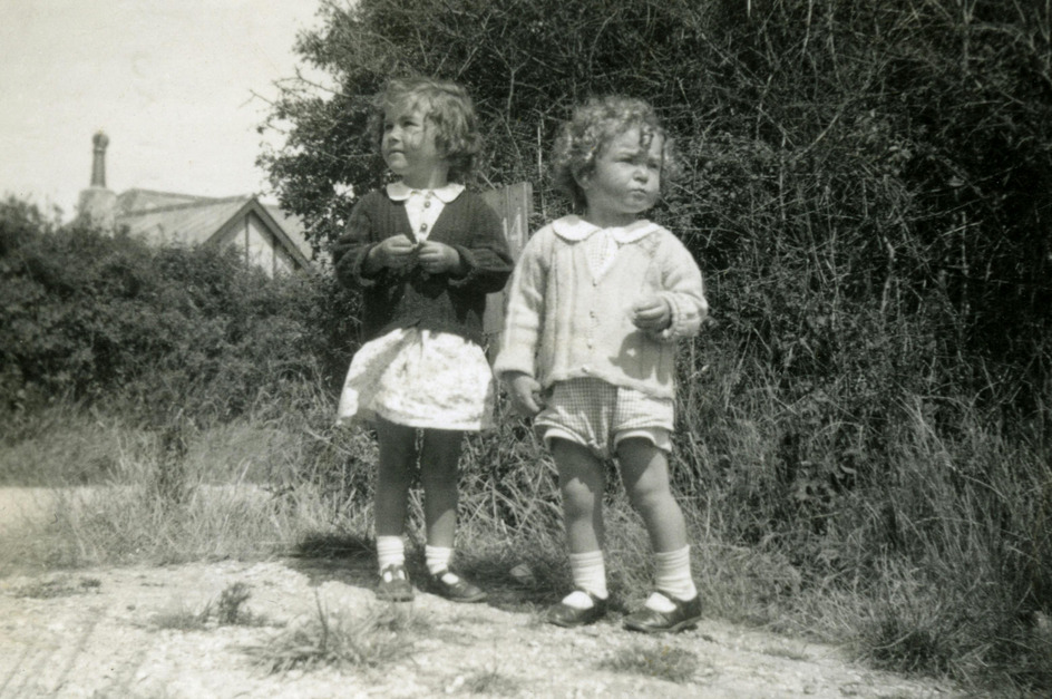 Zwei Kinder stehen auf einem Weg, umgeben von hohen Gräsern und Büschen. Das ältere Kind trägt ein dunkelblaues Oberteil und einen weißen Rock, während das jüngere Kind in einem hellen Strickpullover und kurzen Hosen gekleidet ist. Beide blicken nachdenklich in die Ferne. Das Foto ist in schwarz-weiß.