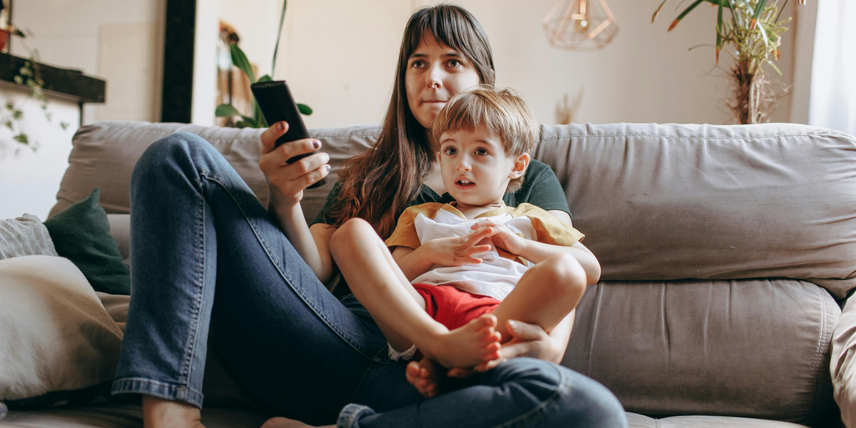 Eine Frau sitzt auf einem Sofa mit einem kleinen Jungen auf ihrem Schoß. Sie hält eine Fernbedienung in der Hand und schaut konzentriert. Der Junge trägt ein T-Shirt und Shorts und sieht interessiert aus.