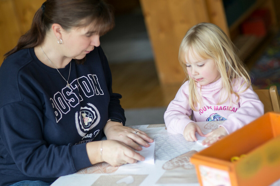 Eine Frau und ein Mädchen sitzen an einem Tisch und basteln. Die Frau zeigt etwas, während das Kind konzentriert mit Papier arbeitet. Auf dem Tisch liegt eine orangefarbene Box und Bastelmaterialien.