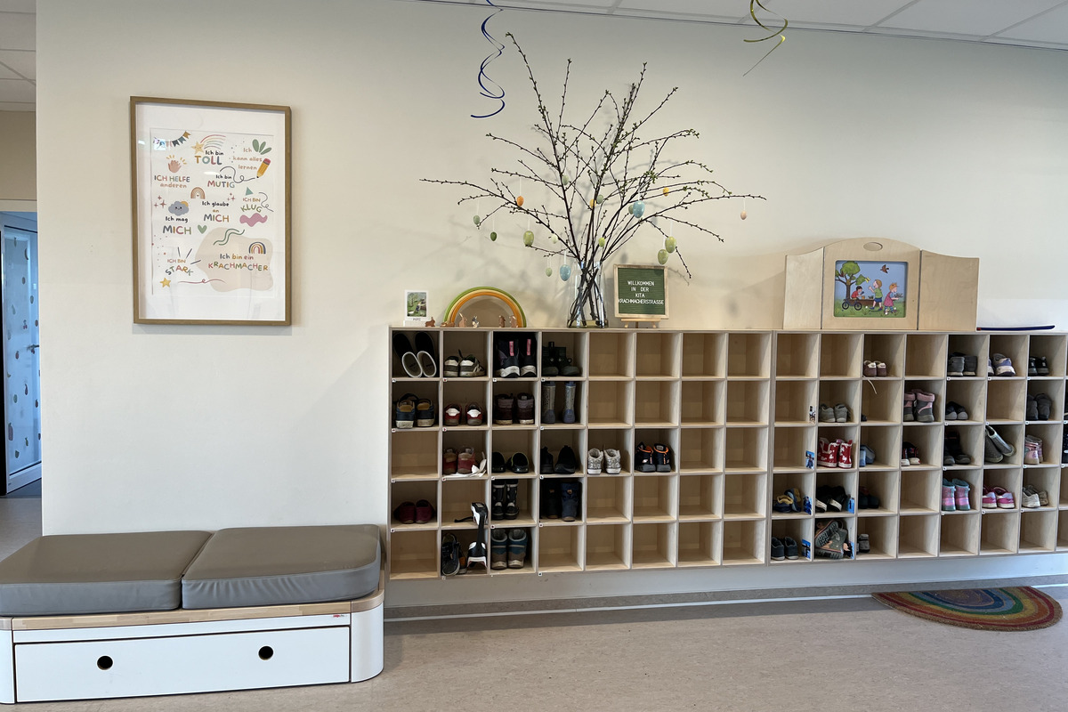 In einem Raum befinden sich Schuhregale mit verschiedenen Schuhen. An der Wand hängt ein Bild mit positiven Sprüchen und ein Baum mit bunten Eiern. Auf einer Ablage steht ein Willkommensschild. Eine auf einem Teppich platzierte Sitzgelegenheit bietet Platz zum Ausruhen.