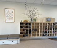 In einem Raum befinden sich Schuhregale mit verschiedenen Schuhen. An der Wand hängt ein Bild mit positiven Sprüchen und ein Baum mit bunten Eiern. Auf einer Ablage steht ein Willkommensschild. Eine auf einem Teppich platzierte Sitzgelegenheit bietet Platz zum Ausruhen.