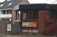 Ein Gebäude mit einem roten Backstein. An der Fassade hängt ein bunter Banner, der eine Veranstaltung ankündigt. D