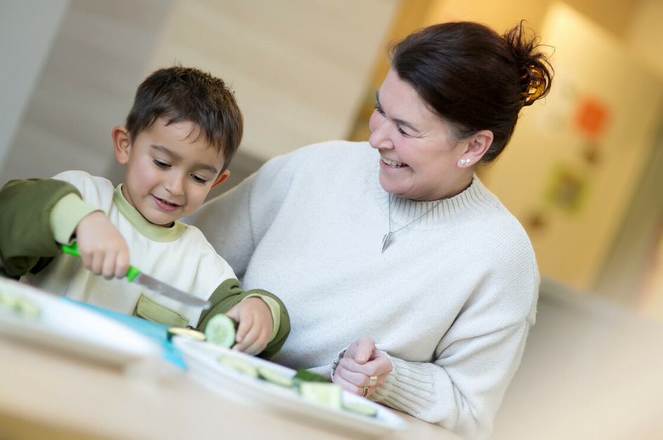 Eine Erzieherin und ein Kind schneiden gemeinsam Gurken. Der Junge lächelt und hält ein Messer, während die Frau ihn beobachtet und lächelt. Auf dem Tisch liegen bereits geschnittene Gurkenscheiben.