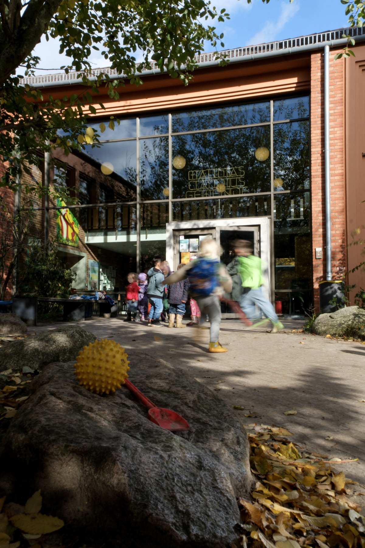 Vor einem Gebäude mit großen Fenstern stehen spielende Kinder. Im Vordergrund liegt ein gelber Ball und eine rote Schaufel auf einem Stein. Herbstblätter bedecken den Boden. 