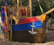 Ein bunt gestaltetes Spielschiff aus Holz mit einem Mast, an dem eine Piratenflagge weht. Auf dem Schiff steht eine figurenartige Figur in Seemannskleidung. Das Schiff hat verschiedene Farben, darunter rot, blau und gelb, und ist von Holzschnitzeln umgeben.