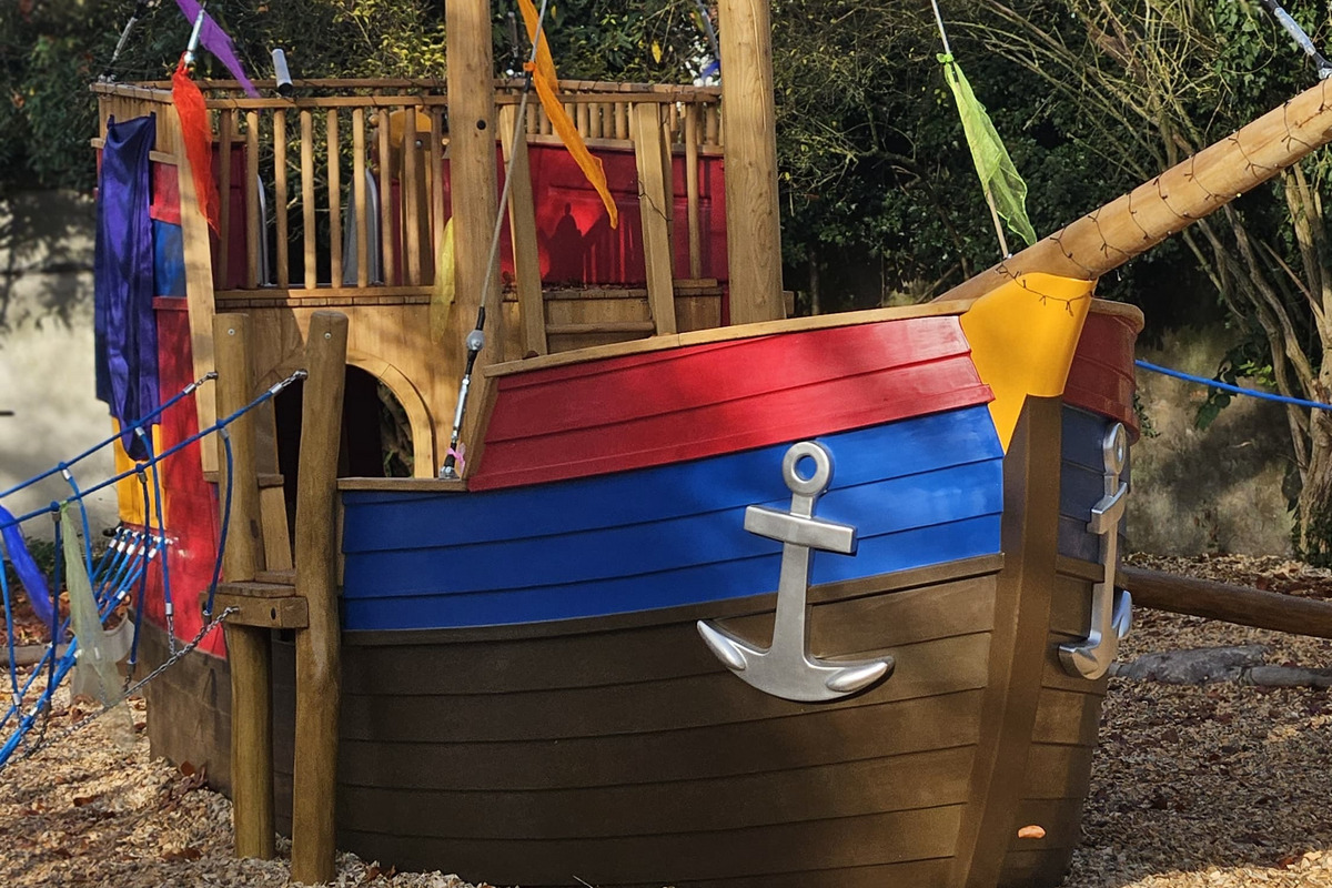 Ein bunt gestaltetes Spielschiff aus Holz mit einem Mast, an dem eine Piratenflagge weht. Auf dem Schiff steht eine figurenartige Figur in Seemannskleidung. Das Schiff hat verschiedene Farben, darunter rot, blau und gelb, und ist von Holzschnitzeln umgeben.