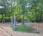 Ein Spielplatz umgeben von Bäumen. Auf der linken Seite befindet sich eine Schaukel mit einem blauen Sitz. 