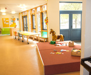 Ein heller Raum mit großen Fenstern, in dem ein Spielbereich zu sehen ist. In der Mitte steht ein Spielpferd und verschiedene Spielmaterialien liegen auf einer gepolsterten Fläche. Es gibt einen Tisch mit Stühlen sowie ein Bücherregal im Hintergrund.
