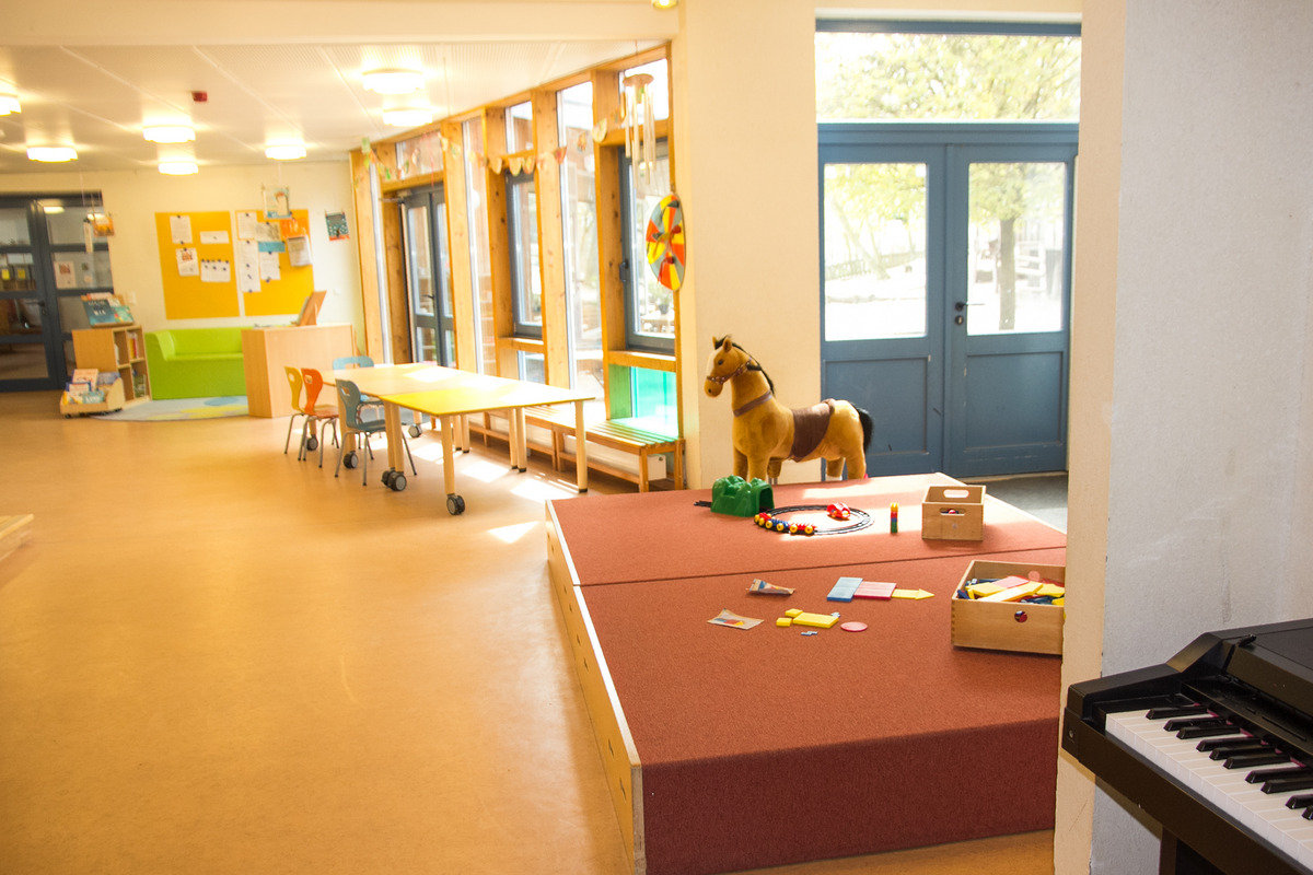 Ein heller Raum mit großen Fenstern, in dem ein Spielbereich zu sehen ist. In der Mitte steht ein Spielpferd und verschiedene Spielmaterialien liegen auf einer gepolsterten Fläche. Es gibt einen Tisch mit Stühlen sowie ein Bücherregal im Hintergrund.