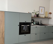 Moderne Küche mit grauen Küchenschränken und einem Holzelement. Eine integrierte schwarze Backofen- und Kochstelle ist sichtbar. Auf dem Regal darüber stehen Pflanzen, eine Kaffeemaschine und eine kleine Lampe. Ein Handtuch hängt vor dem Ofen.