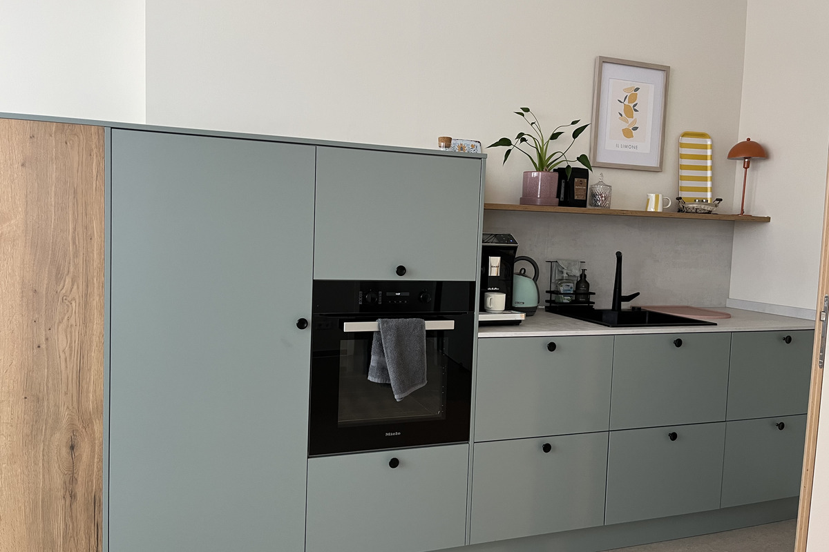 Moderne Küche mit grauen Küchenschränken und einem Holzelement. Eine integrierte schwarze Backofen- und Kochstelle ist sichtbar. Auf dem Regal darüber stehen Pflanzen, eine Kaffeemaschine und eine kleine Lampe. Ein Handtuch hängt vor dem Ofen.
