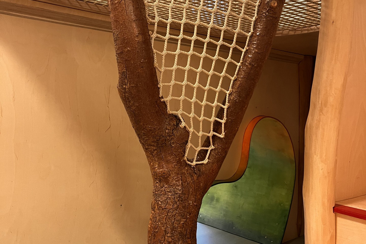 Ein Innenbereich mit einem Baumstamm, der Teil einer Spielstruktur ist. Über dem Stamm ist ein Netz gespannt, das als Sicherheitsvorrichtung dient. Im Hintergrund befindet sich eine farbige, geschwungene Wandöffnung. Der Boden besteht aus einem glatten, grauen Material.