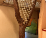 Ein Innenbereich mit einem Baumstamm, der Teil einer Spielstruktur ist. Über dem Stamm ist ein Netz gespannt, das als Sicherheitsvorrichtung dient. Im Hintergrund befindet sich eine farbige, geschwungene Wandöffnung. Der Boden besteht aus einem glatten, grauen Material.