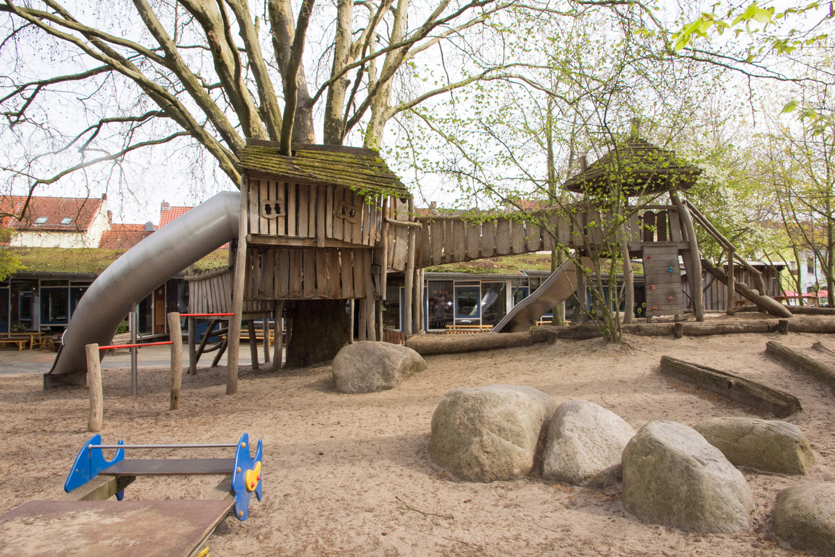 Ein Spielplatz mit einem großen Baum, der ein Holzhäuschen beherbergt. Rutsche und Kletterelemente sind vorhanden. Der Boden ist mit Sand und großen Steinen bedeckt, umgeben von einigen Bäumen und einem Gebäude im Hintergrund.