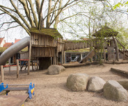 Ein Spielplatz mit einem großen Baum, der ein Holzhäuschen beherbergt. Rutsche und Kletterelemente sind vorhanden. Der Boden ist mit Sand und großen Steinen bedeckt, umgeben von einigen Bäumen und einem Gebäude im Hintergrund.