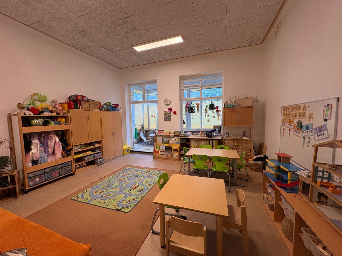 Ein heller Klassenraum mit Tischen und Stühlen in verschiedenen Farben. Eine gemütliche Ecke mit einem Teppich und einem Regal voller Spielzeuge. Fenster lassen Tageslicht herein, und im Hintergrund sind Regale mit Lehrmaterialien sichtbar.