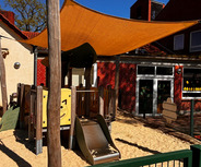 Ein Spielplatz mit einer Rutsche, umgeben von Sand, befindet sich unter einem orangefarbenen Sonnensegel. Im Hintergrund sind Teile eines Gebäudes zu sehen, das hinter einem Zaun steht. Der Spielplatz zeigt bunte, tierähnliche Elemente.