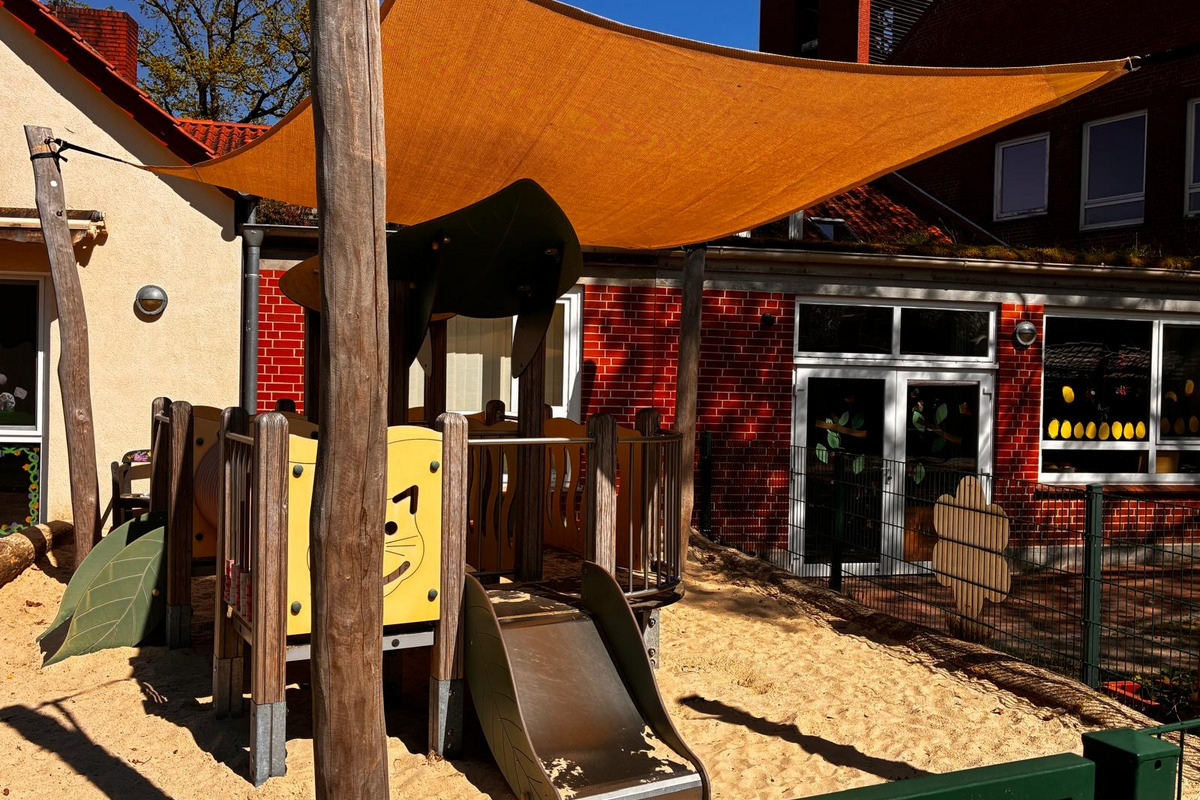 Ein Spielplatz mit einer Rutsche, umgeben von Sand, befindet sich unter einem orangefarbenen Sonnensegel. Im Hintergrund sind Teile eines Gebäudes zu sehen, das hinter einem Zaun steht. Der Spielplatz zeigt bunte, tierähnliche Elemente.