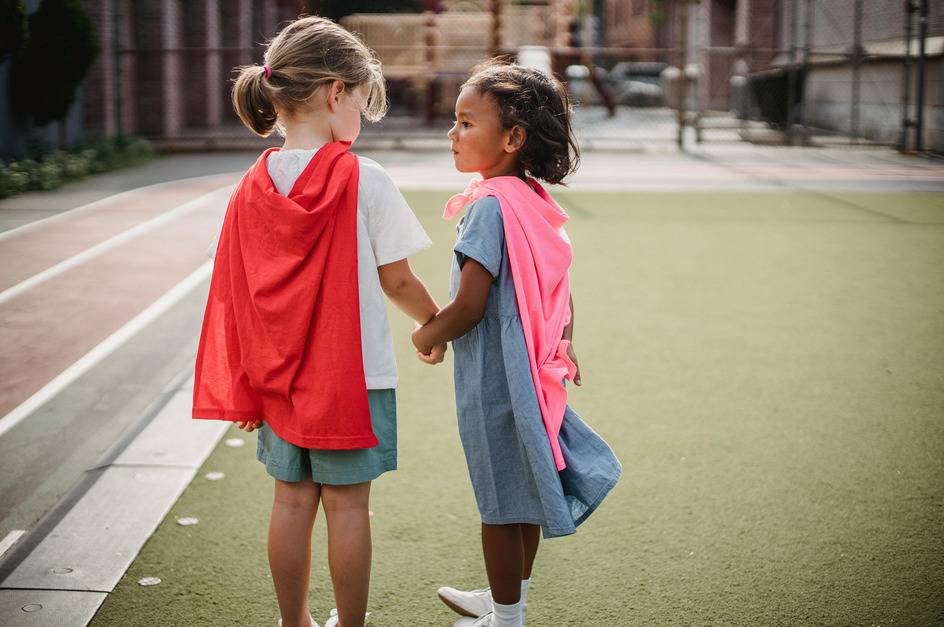 Zwei kleine Mädchen stehen auf einem Sportplatz. Eines trägt einen roten Umhang, das andere einen pinken. Sie halten sich an den Händen und schauen sich an.
