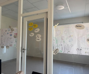 In einem Büro sind mehrere großformatige Zeichnungen und Plakate an Fenstern befestigt. Die Zeichnungen enthalten bunte Kritzeleien und Überschriften, die verschiedene Themen oder Ideen beschreiben.