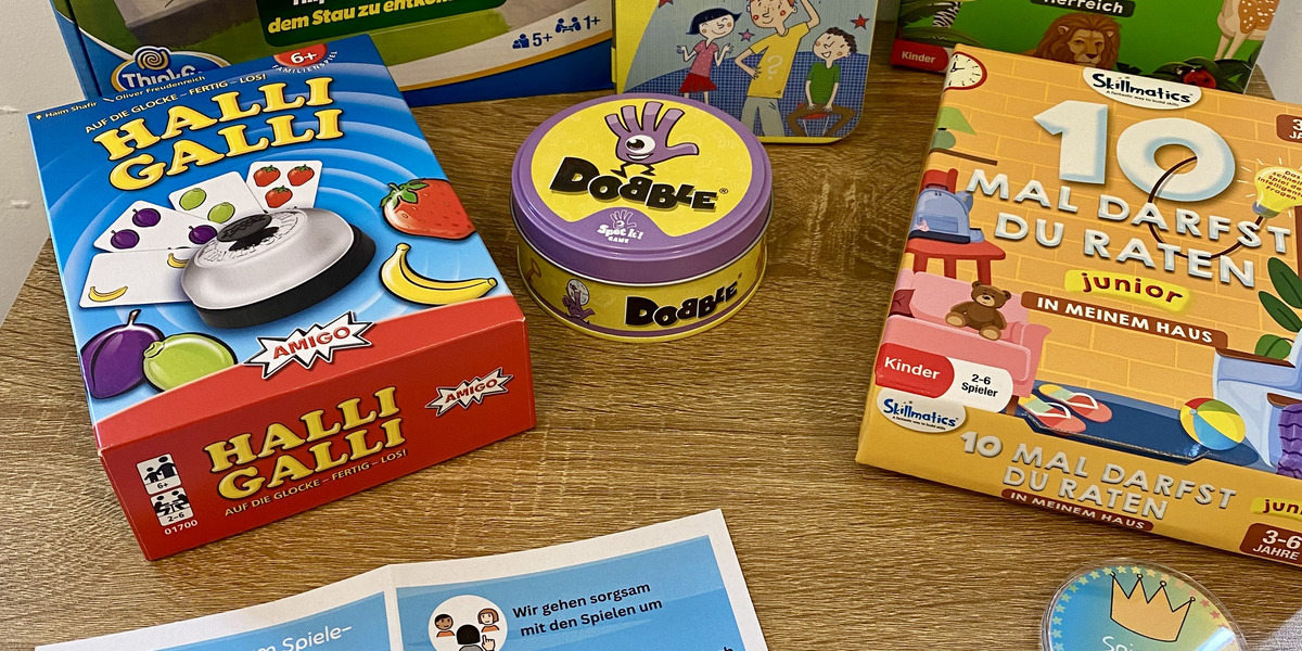 Auf einem Tisch liegen verschiedene Gesellschaftsspiele für Kinder. Dazu gehören "Rush Hour Junior", "Halli Galli", "Dobble" und "Wer bin ich?". Außerdem befindet sich ein Flyer mit Hinweisen zur Spielnutzung und Belohnungen für das Spielen.