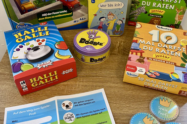 Auf einem Tisch liegen verschiedene Gesellschaftsspiele für Kinder. Dazu gehören "Rush Hour Junior", "Halli Galli", "Dobble" und "Wer bin ich?". Außerdem befindet sich ein Flyer mit Hinweisen zur Spielnutzung und Belohnungen für das Spielen.